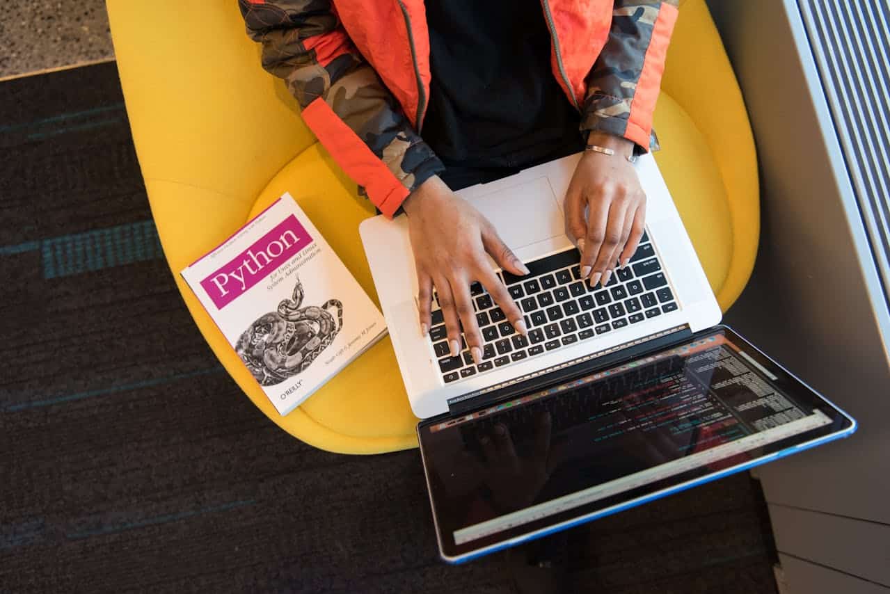 Persona che programma in Python su laptop con libro O'Reilly visibile, seduta su poltrona gialla — concetto di sviluppo software e coding.